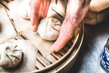Nahaufnahme von Händen, die rohe Dumplings zum Dämpfen in einen Bambuskorb setzen