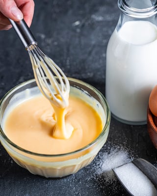 Weiße Schokocreme in kleiner Schüssel mit Schneebesen, daneben stehen Eier und Milch