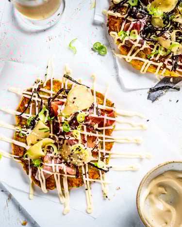 Okonomiyaki – Japanischer Pfannkuchen m,it Möhren, Weißkohl, Algen und Ingwer. Fotografiert von oben, mit Mayonnaise und Sesam garniert, auf weißem Untergrund.