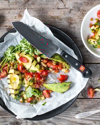 Gegrillte Avocado mit Tomatensalsa und Rucola auf Teller verteilt mit Besteck auf Holzuntergrund.