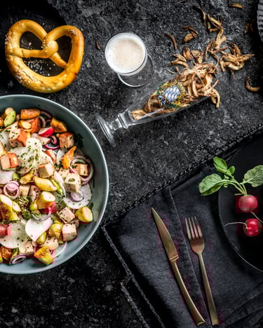 Ein Teller mit bayerischem Brotsalat steht auf dunklem Untergrund neben einem ganzen gegrillten Brathähnchen, Brezeln, Radieschen und einem umgestürzten Bierglas mit Röstzwiebeln