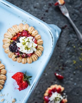 Schnelle Tartelettes mit Beerenfüllung und einer Sahnehaube mit Pistazien auf einem Backblech und eine angebissene Erdbeere