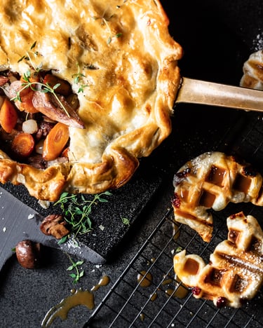 Eine Pfanne mit Pot Pie, bei dem durch die angebrochene Blätterteighaube die Pilz-Füllung sichtbar wird, und daneben Croffles mit Karamellsauce auf einem dunklen Untergrund