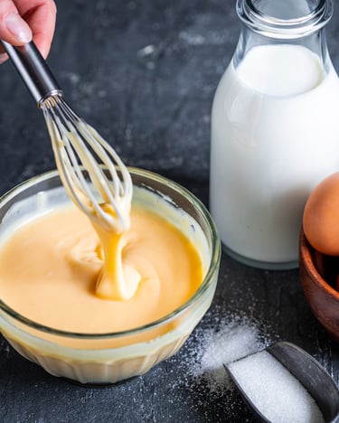 Crema pasticcera mit weißer Schokolade in Schüssel. Daneben Milchkanne und Eier.