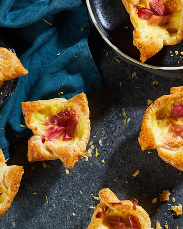 Mehrere Tartelettes liegen auf grauem Untergrund. Daneben im Anschnitt eine Schüssel mit Rhabarber