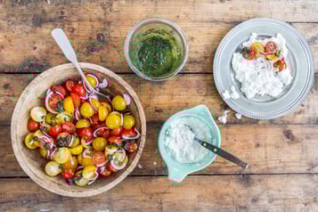 In einer großen Holzschale ist Tomatensalat in dem ein Esslöffel liegt. Daneben eine Schale mit Hüttenkäse und einem Löffel, eine Schale mit Basilikumpaste und einem Teller auf dem Hüttenkäse mit Tomatensalat bestückt ist.