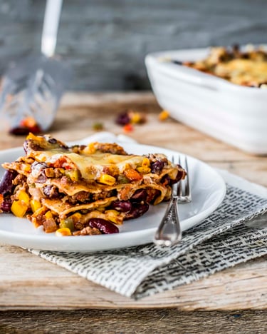 Auf einem weißen Teller ein Stück Lasagne mit Kidneybohnen und Mais. Im Hintergrund eine Auflaufform mit Lasagne