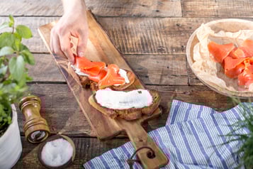 Zwei Graubrotscheiben mit Frischkäse werden mit Räucherlachs belegt. Die Brote liegen auf einem Holzbrett, im Bildrand Kräuter, eine liegende Pfeffermühle eine Schale mit Salz und eine große Schale mit Räucherlachs.