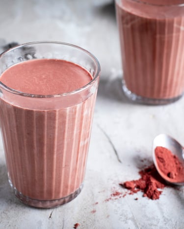 Zwei Gläser mit hellbraunem Bananen-Smoothie neben Löffel mit Kakaopulver auf hellgrauem Tuch