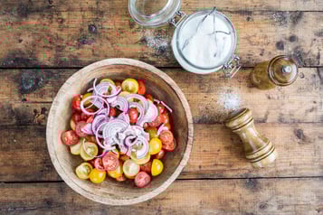 In einer großen Holzschüssel geschnittene Cherrytomaten und Zwiebeln mit Vanillezucker überstreut. Daneben ein Glas mit Vanillezucker und Pfeffermühlen