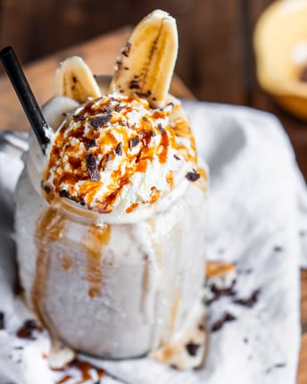 Ein großes Glas mit cremigem Peanutbutter-Banana-Milchshake, das mit einer Kugel  Stracciatella-Eis, Sprühsahne, Karamellsauce und einer halbierten Banane getoppt ist.