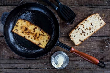 Eine Scheibe Rosinenbrot wird in einer öligen Pfanne angeröstet. Daneben liegen Pfannenwender und eine Scheibe ungeröstetes Rosinenbrot sowie eine kleine Schale mit Butter.