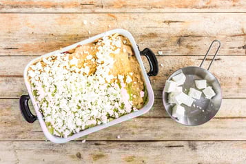 Gebröselter Feta auf einer Lasagne, daneben eine Stahlschüssel mit Feta