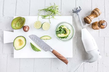 Auf einem Schneidebrett befindet sich eine Avocado und ein Pürierstab.