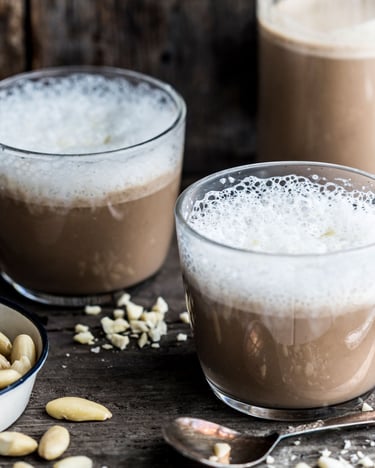 Drei Gläser mit Espresso-Smoothie und Mandelmilchschaum neben Mandeln auf Holzgrund