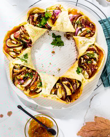 Auf einem Kuchenteller liegt ein Quiche-Ring mit Feigen und Ziegenkäse, darunter ein rundes Küchengitter. Links im Bild ein Glas mit Fruchtaufstrich der Marke St. Dalfour und eine kleine Schale mit Fruchtaufstrich und einem Löffel.