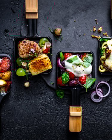 Vier bunt gefüllte Raclette-Pfännchen mit Gemüse, Fleisch und Käse