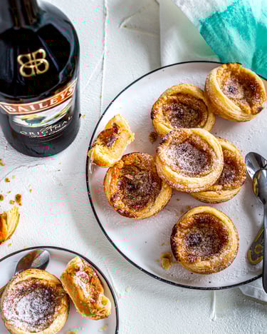 Mehrere Pasteis de Nata mit Baileys auf weißem Teller mit türkiser Stoffserviette.