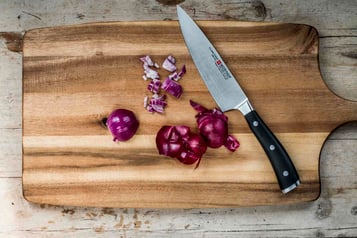 Gewürfelte rote Zwiebel auf einem Holzbrett. Daneben eine Zwiebelhälfte, ein großes Küchenmesser sowie Zwiebelschale