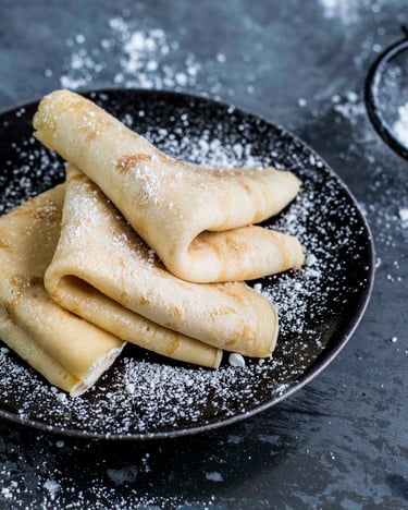 Auf einem schwarzen Teller liegen drei gefaltete Pfannkuchen, daneben ein Sieb mit Puderzucker