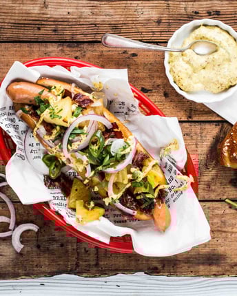 Laugen-Hotdog in einem roten Servierkörbchen mit Würstchen, Bacon, Zwiebeln und Ananas mit einem Schälchen Dijonnaise auf Holzuntergrund.