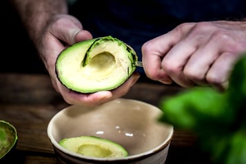 Die Avocado wird in eine Schüssel gegeben, im Vordergrund steht ein Basilikum
