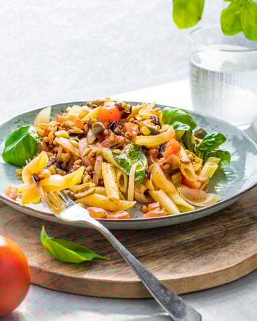 Penne mit Caponata auf Holzbrett, daneben liegt eine Tomate und eine Gabel