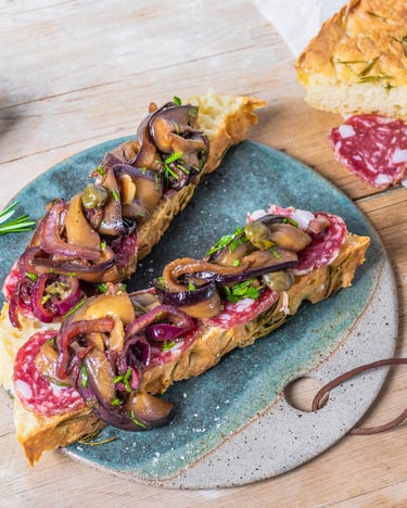 Zwei Scheiben Focaccia mit Aubergine und Salami belegt, daneben liegt Rosmarin