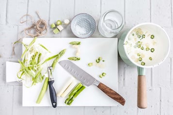 Auf einem Schneidebrett befindet sich Spargel, ein Sparschäler und ein Schneidemesser.