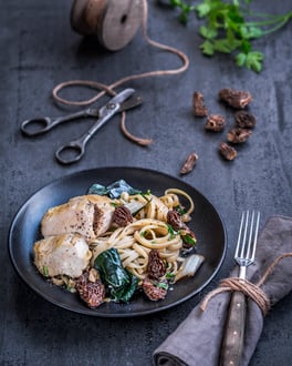 Linguine mit Hähnchenbrust, Mangold und Morcheln angerichtet auf dunklem Teller und dunklem Holzuntergrund. Daneben eine Gabel mit Kordel an Stoffserviette gewickelt.