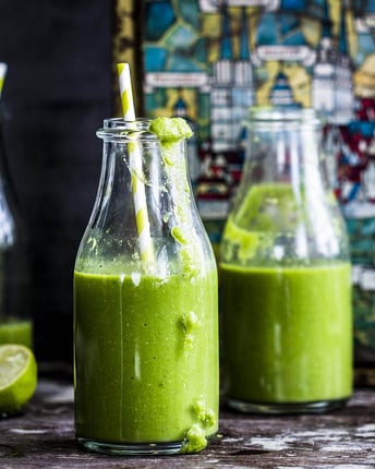 Drei kleine Glasflaschen mit grünem Smoothie und Strohalmen auf dunklem Holzgrund