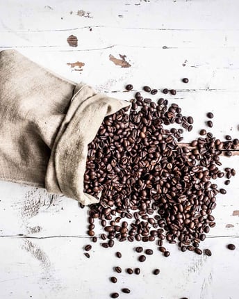 Ein umgefallener Kaffeesack mit herausfallenden gerösteten Kaffeebohnen von oben auf einem hellen Holzuntergrund