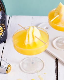 Zwei Gläser mit sonnengelbem Flirtini und Ananas-Stücken am Spieß auf einem weißen Holztisch vor türkisem Hintergrund
