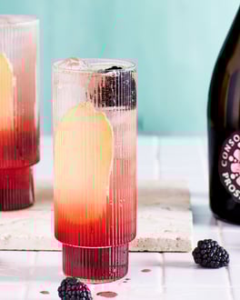 Zwei Gläser mit Brombeer Mule mit Prosecco Rosé. An der Glasseite ist eine längs geschnittene Ingwerscheibe zu sehen, oben schwimmen Eiswürfel und frische Brombeeren sind in Glas und auf dem weißen Fliesenuntergrund verteilt.