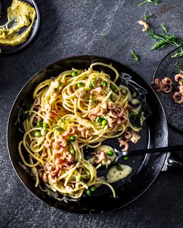 Eine Pfanne mit Spaghetti in Weißweinsoße, dekoriert mit Nordseekrabben ist von oben fotografiert. Daneben liegt ein Senfglas und ein paar Krabben.