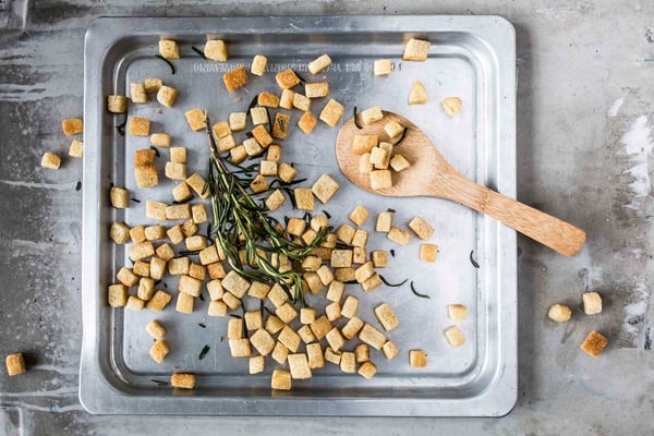 Croutons liegen verteilt auf einem grauen Blech. Darauf Rosmarin-Stengel