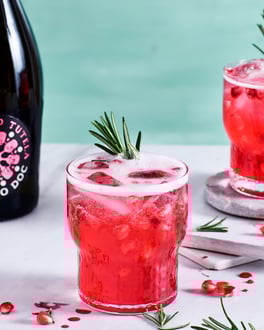 Zwei Gläser mit rotem Granatapfel-Mimosa, Eiswürfeln, frischen Granatapfelkernen und Rosmarin vor einem hellen Holzuntergrund und türkischem Hintergrund.