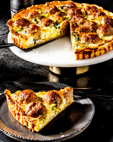 Eine angeschnittene Quiche mit Hackbällchen und cremiger Ei-Sahne-Masse steht auf einer weißen Tortenplatte vor dunklem Hintergrund neben einem schwarzen Steller mit einem Stück Hackbällchen-Quiche