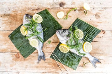 2 Fische liegen mit Dill und Zitronenscheiben auf Bananenblättern