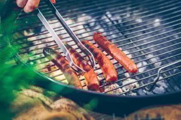 Auf einem Grillrost liegen 4 Lammbratwürste, von denen eines gerade mit einer Grillzange umgedreht wird.