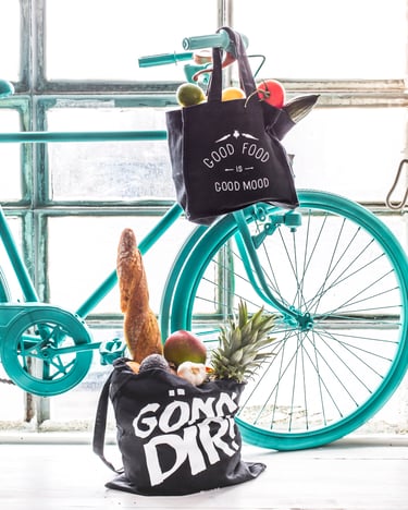 Zwei schwarze Jutebeutel, die mit einem Baguette und frischem Obst und Gemüse gefüllt sind, stehen vor einem türkisen Fahrrad auf dem Boden