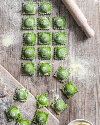 Rohe grüne Ravioli auf Holzuntergrund, daneben liegt ein Nudelholz