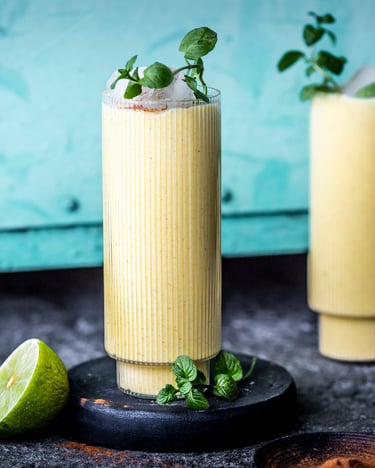 Frontal fotografiert sieht man zwei hohe Gläser, gefüllt mit Mango Lassi und dekoriert mit Minzblättern vor einer Türkisen Wand