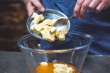 Eine Person gibt mit einem Löffel Butterwürfel in eine Glasschüssel.