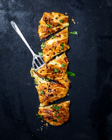 Ein angeschnittener goldbraun gebackener Blätterteig-Zopf mit Tomate-Mozzarella-Füllung und Topping aus Basilikum und Pinienkernen vor einem dunklen Untergrund