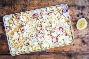 Auf Holzuntergrund liegt ein Backblech mit Blumenkohl und Zwiebeln,daneben eine Zitrone