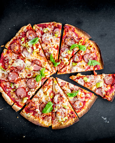 Eine runde Pizza mit krossem Rand und Belag aus Tomatensauce, Cabanossi, Frühlingszwiebeln und Käse liegt geschnitten auf schwarzem Untergrund.