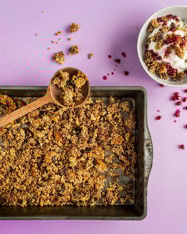 Salted Caramel Granola mit Quinoa auf einem tiefen Backblech. Darüber liegt quer ein großer Holzlöffel mit Granola-Stückchen. Rechts oben im Bild eine Schüssel mit Joghurt und Granola.