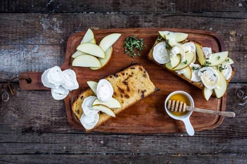 Auf einem Holzbrett liegen zwei Scheiben Rosinenbrot. Das eine ist komplett mit Apfelscheiben, Ziegenkäse und Kräutern belegt. Das andere nur zur Hälfte. Daneben liegen Apfelscheibe und Ziegenkäsescheiben sowie eine kleine Schale mit Honig.