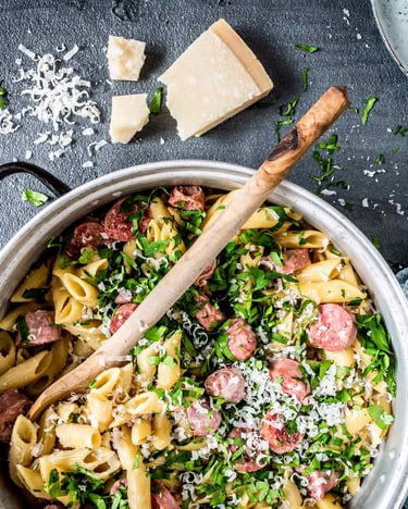 Topf mit Penne, Salsiccia-Stücken, Petersilie und Parmesan auf grauem Grund neben Parmesanstück, Gabeln und Tuch